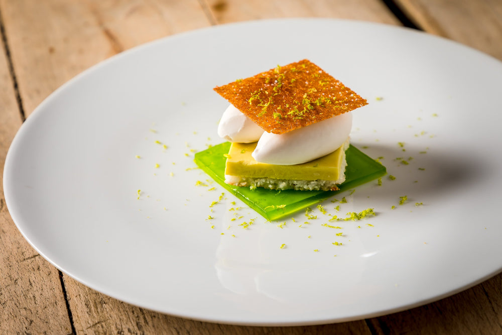 Un dessert sophistiqué avec une présentation en couches, surmonté d'une fine croustille et de zeste de citron vert, servi sur une assiette blanche.