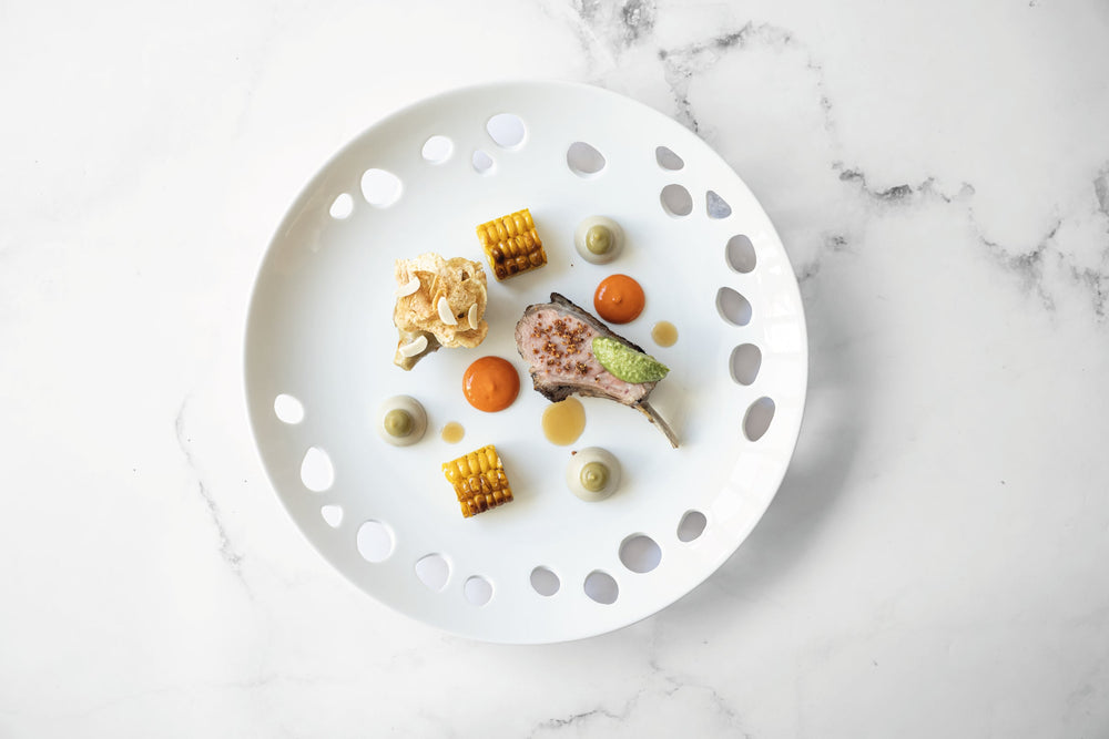 Assiette gastronomique élégamment dressée avec un carré d'agneau, des cubes de maïs grillé, des purées colorées et des croustillants, sur une assiette blanche ajourée.