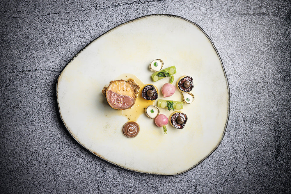Présentation artistique d'un plat gastronomique comprenant une pièce de viande tendre accompagnée de légumes soigneusement disposés et de sauces élégantes, le tout sur une assiette texturée.