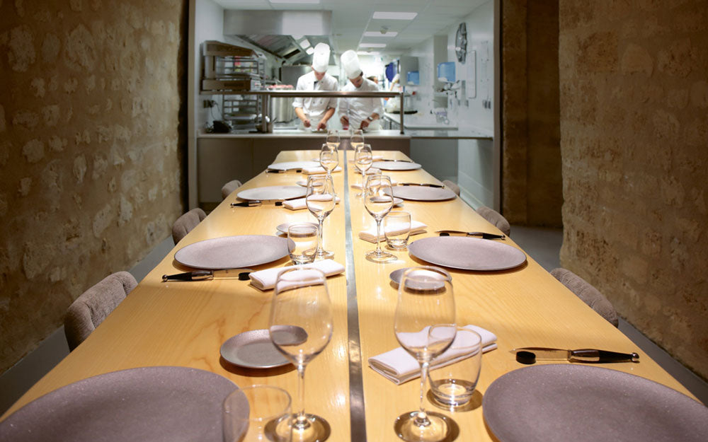 Table d'hôtes élégamment dressée avec vue sur une cuisine ouverte où des chefs en tenue blanche préparent des plats, offrant une expérience culinaire immersive et conviviale.