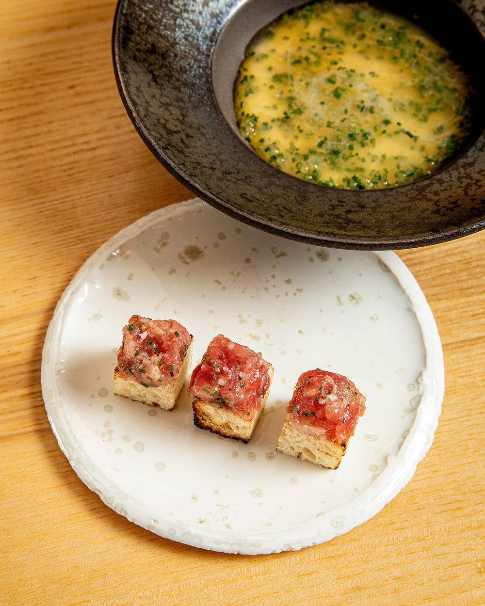 Trois morceaux de tartare de bœuf sur des cubes de pain grillé, accompagnés d'une soupe présentée dans un bol noir.