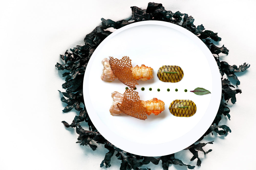 Assiette gastronomique avec des langoustines et des légumes finement décorés, entourée de morceaux de légumes grillés, sur un fond blanc.