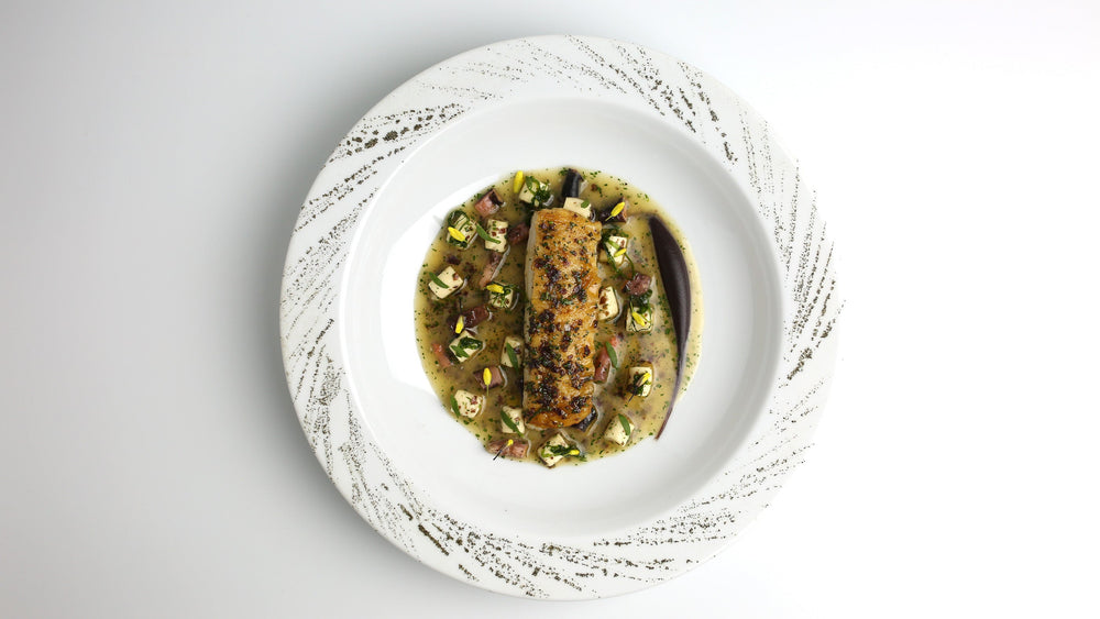 Un plat gastronomique composé d'un filet de poisson parfaitement cuit, servi sur un lit de légumes et accompagné d'une sauce raffinée, le tout présenté dans une assiette blanche ornée de motifs élégants.