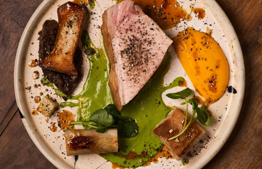 Assiette gastronomique avec un morceau de viande rosée, des purées de légumes vertes et oranges, des champignons et des os à moelle, le tout joliment disposé avec des herbes fraîches.